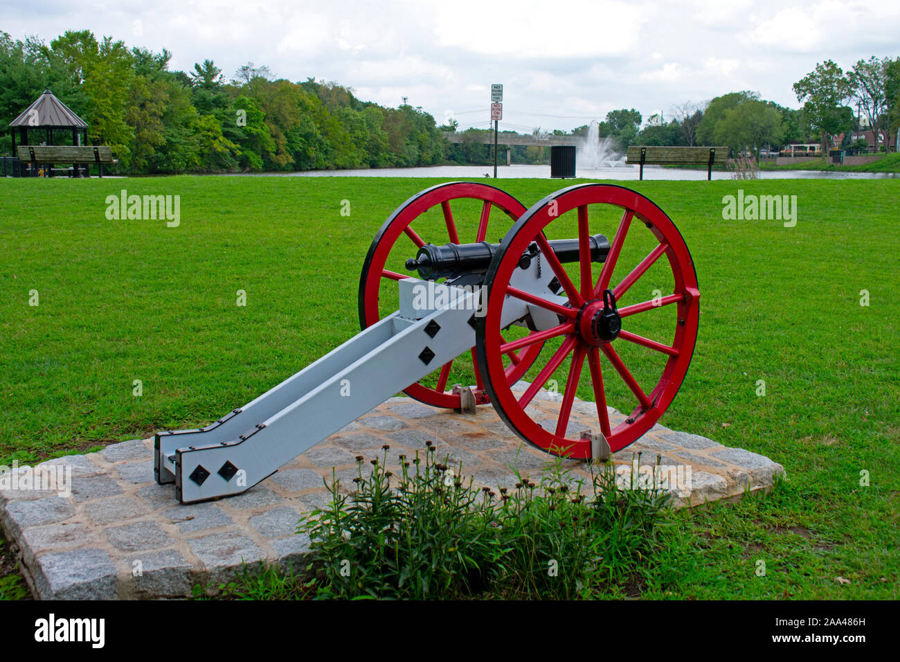 American Revolutionary War cannon with red wheels and a black barrel on display in front of a water fountain at Columbus Park, Piscataway, New Jersey, Stock Photo