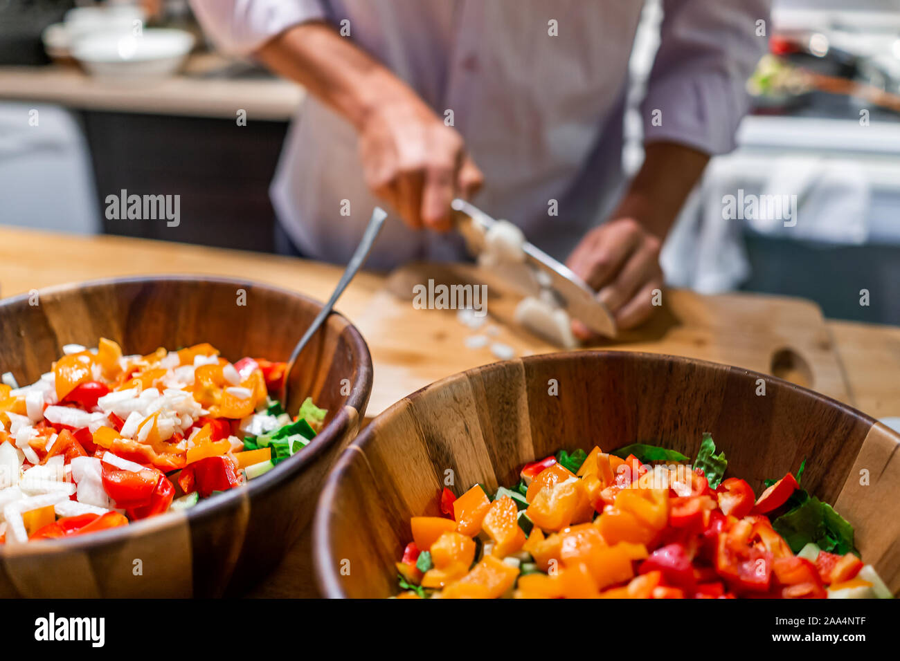 Man chopping onions on cutting board table in background with two large bowls of fresh vegan salad with red orange bell peppers in kitchen Stock Photo