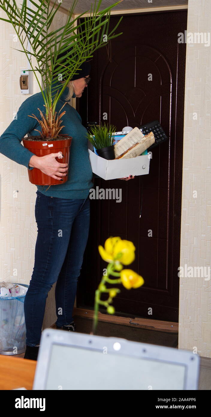 Sad adult woman fired from work is standing near the office door with a box of belongings and a beloved palm tree. Stock Photo