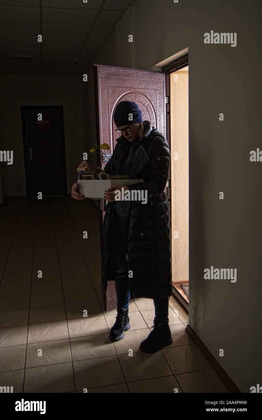 Sad adult woman fired from work is standing near the office door with a box of belongings. Stock Photo