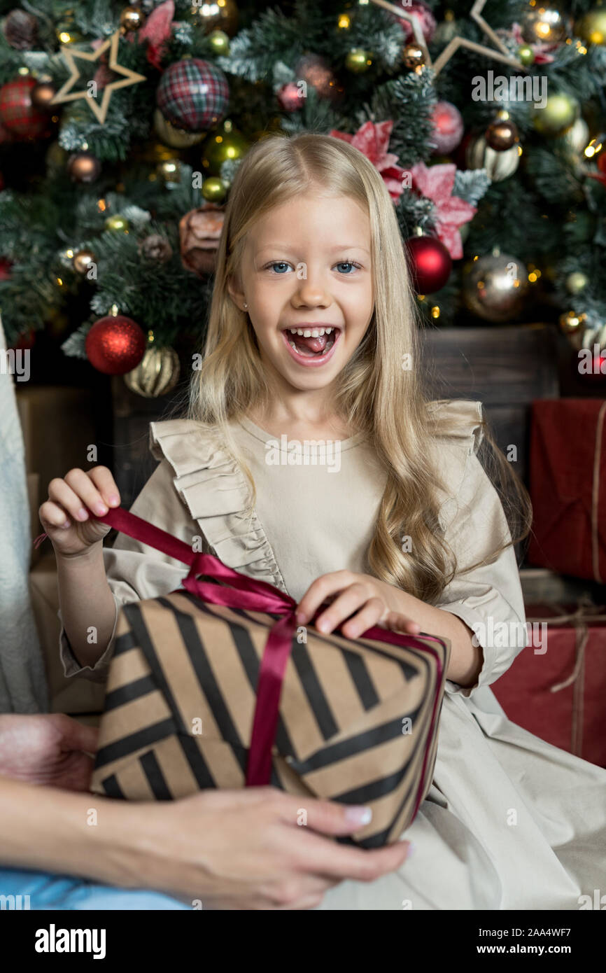 Smiling girl sitting in front of a Christmas tree unwrapping a gift Stock Photo