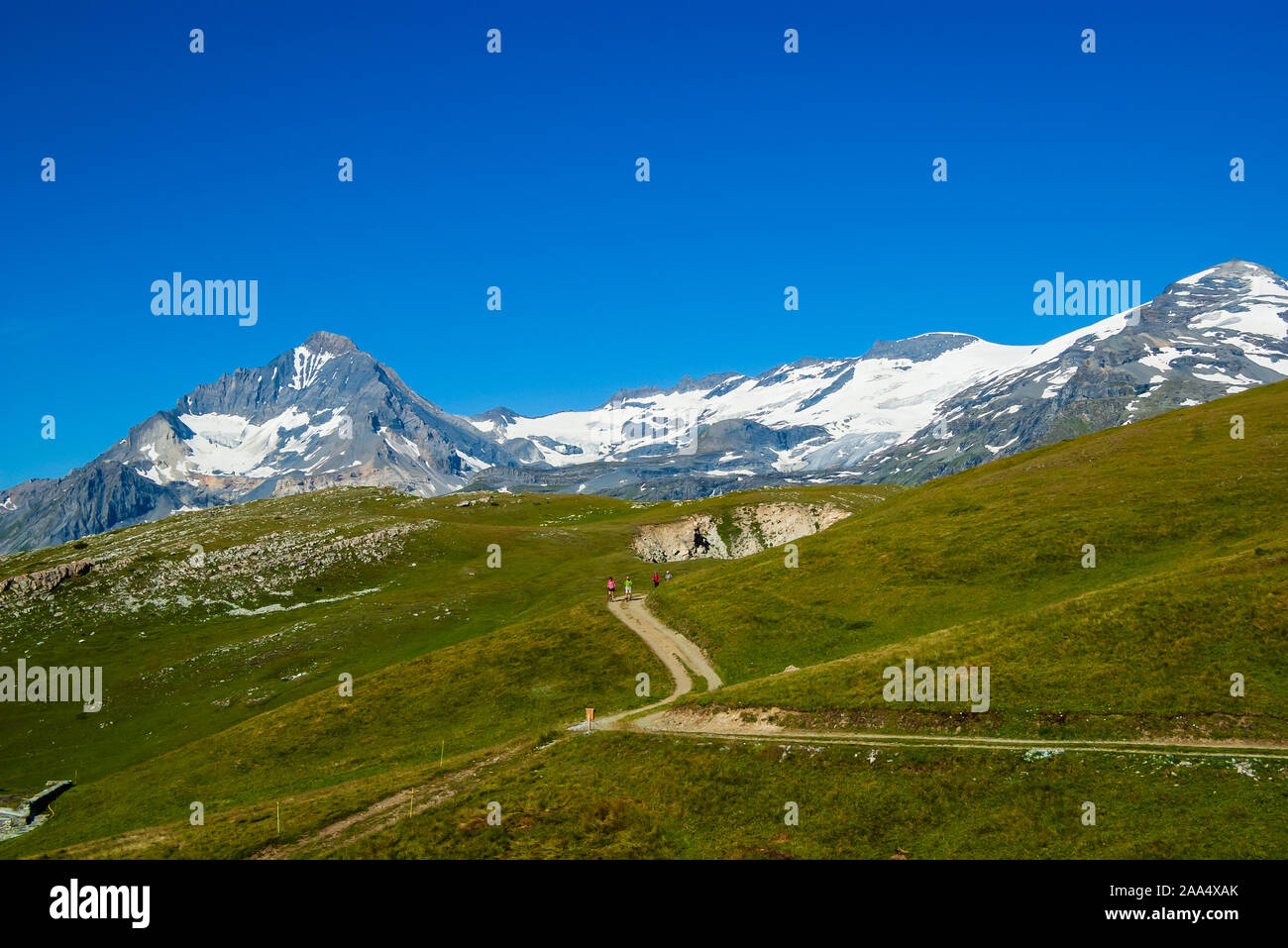 Summer Alpine landscape in French Alps. Hiking trail with wondering tourists in the far distance. Green alpine pastures and mountain with snowy slopes Stock Photo