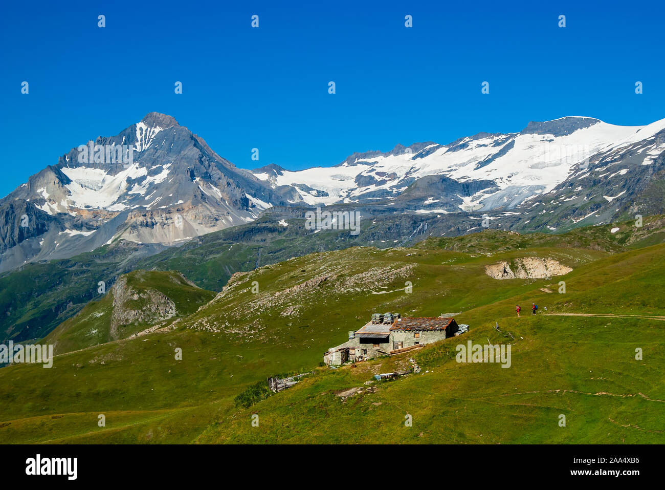 Summer Alpine landscape. Stone farm building high above a green valley in the Vanoise National Park,  a mountain with snowy slopes and a blue sky. Stock Photo