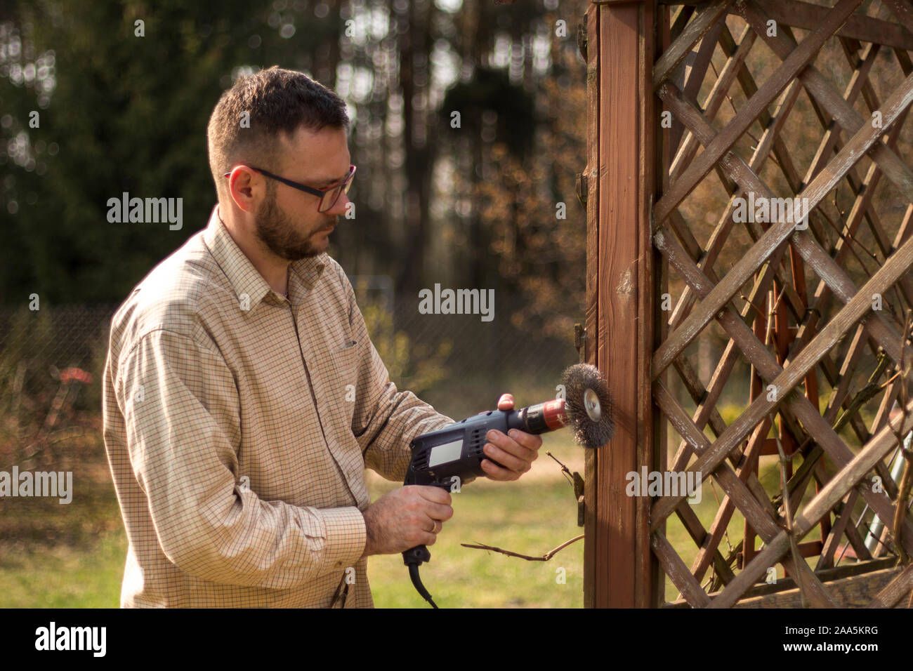 Man with glasses and a flannel shirt during spring gardening. Renovation and maintenance of garden pergolas. Removing old paint from wooden elements. Stock Photo