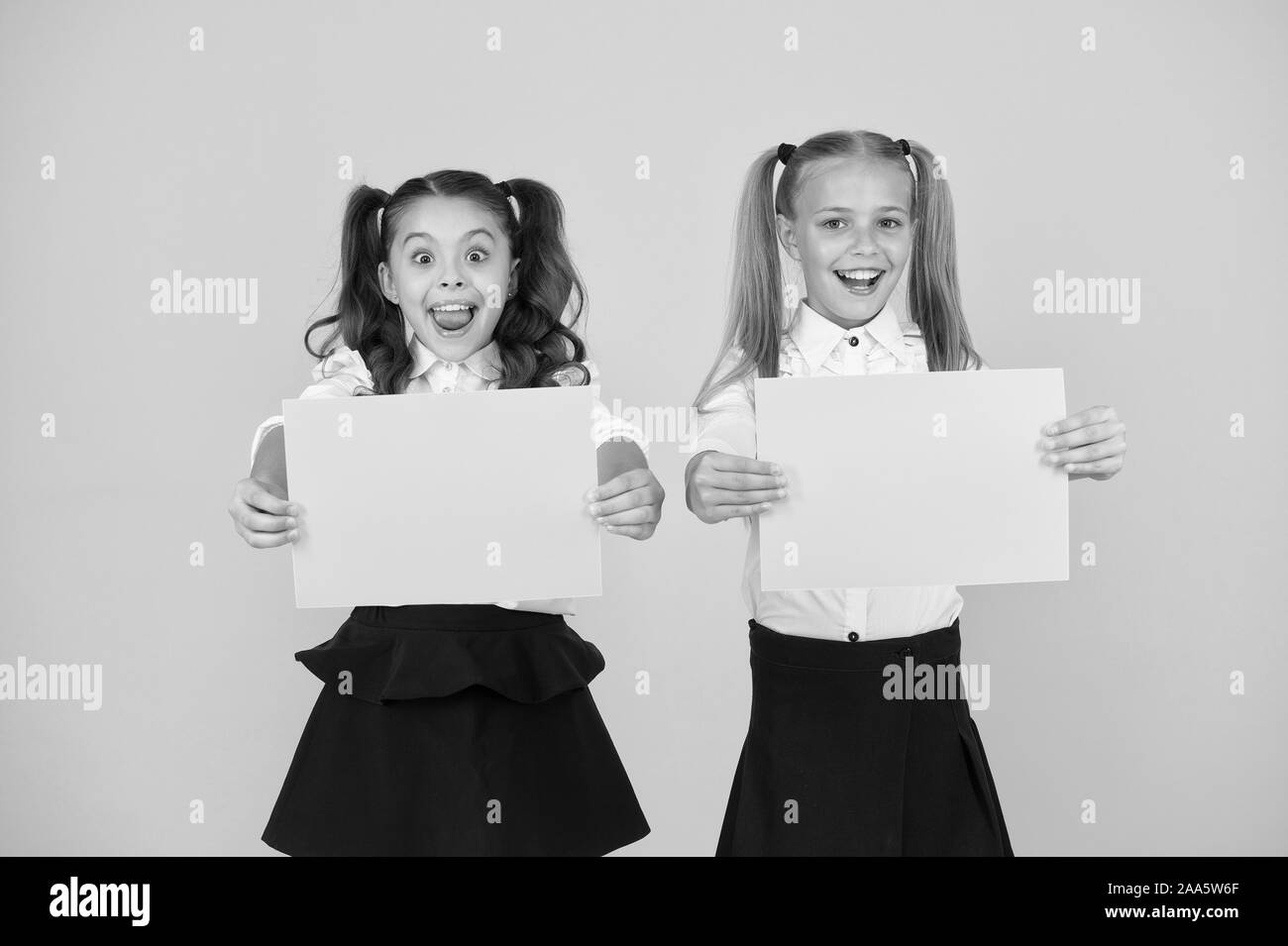 Advertising idea for school. Happy girls holding paper sheets for certain idea on yellow background. Small cute kids smiling of genius idea. Little children with empty paper for your idea, copy space. Stock Photo