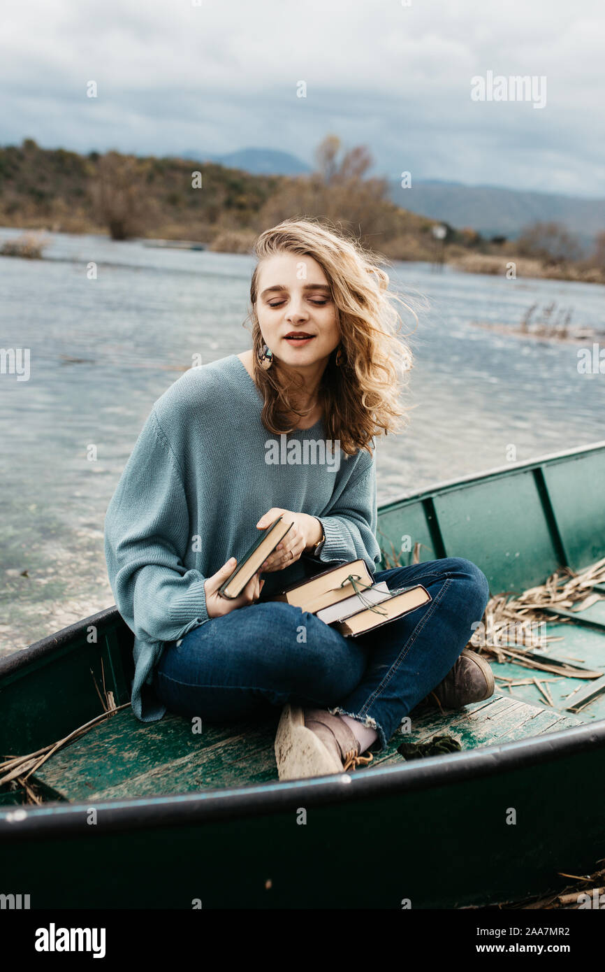 Portrait of young beautiful woman sitting on a boat and reading a book. She is bookworm and she choose between few books. Stock Photo