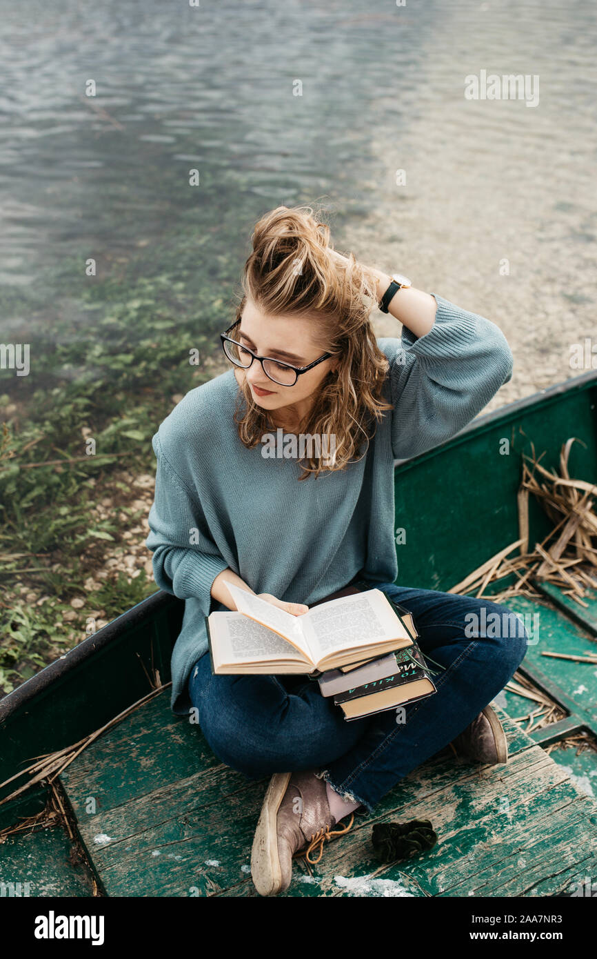 Portrait of young beautiful woman sitting on a boat and reading a book. She is bookworm and she choose between few books. Stock Photo