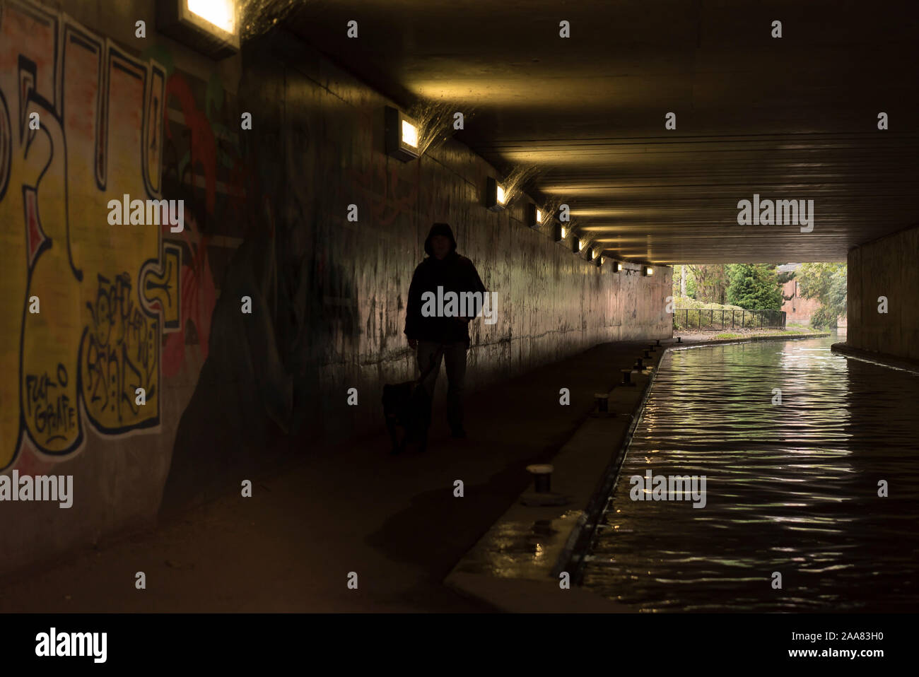 An isolated, hooded, solitary figure walking by an urban canal in dark, UK pedestrian underpass, passing urban graffiti artwork sprayed on walls Stock Photo