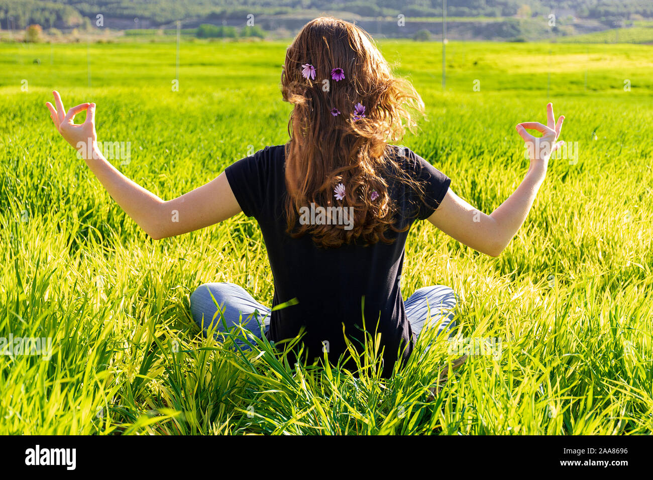 Young caucasian woman red-haired with freckles, resting on a green field at spring sunset, sitting in a yoga position. Meditation, mindfulness, relax Stock Photo