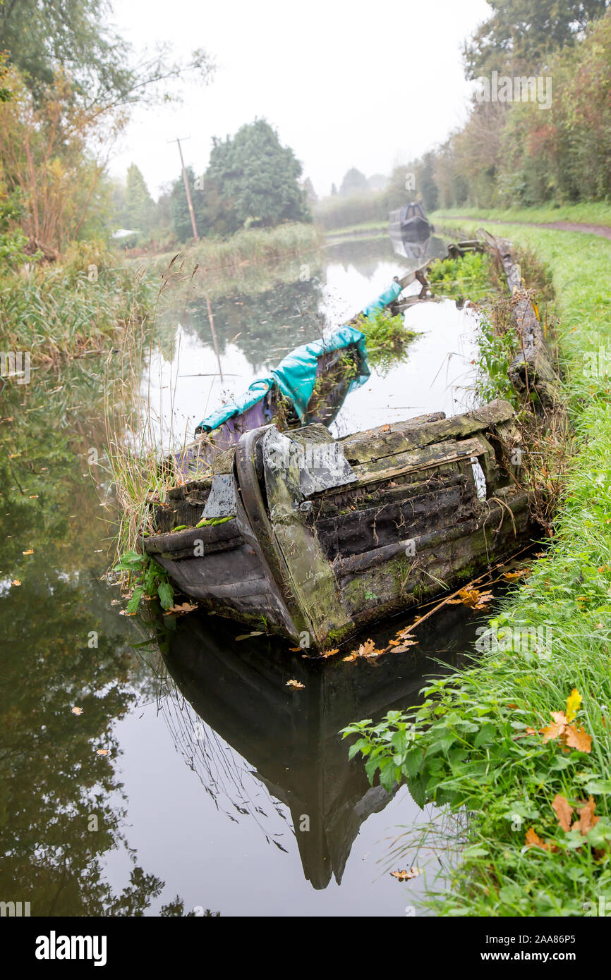 Portrait close up of narrowboat wreckage abandoned in UK canal water on a misty, autumn morning - forgotten, wood rotting & weeds growing inside. Stock Photo