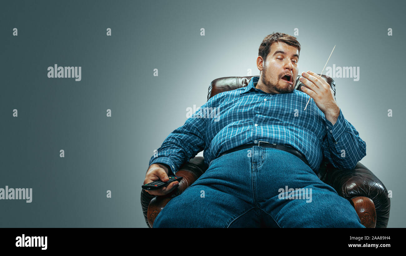 Portrait of fat caucasian man wearing jeanse and whirt sitting in a brown armchair isolated on gradient grey background. Emotional watching TV and changing channels, laughting. Overweight, carefree. Stock Photo