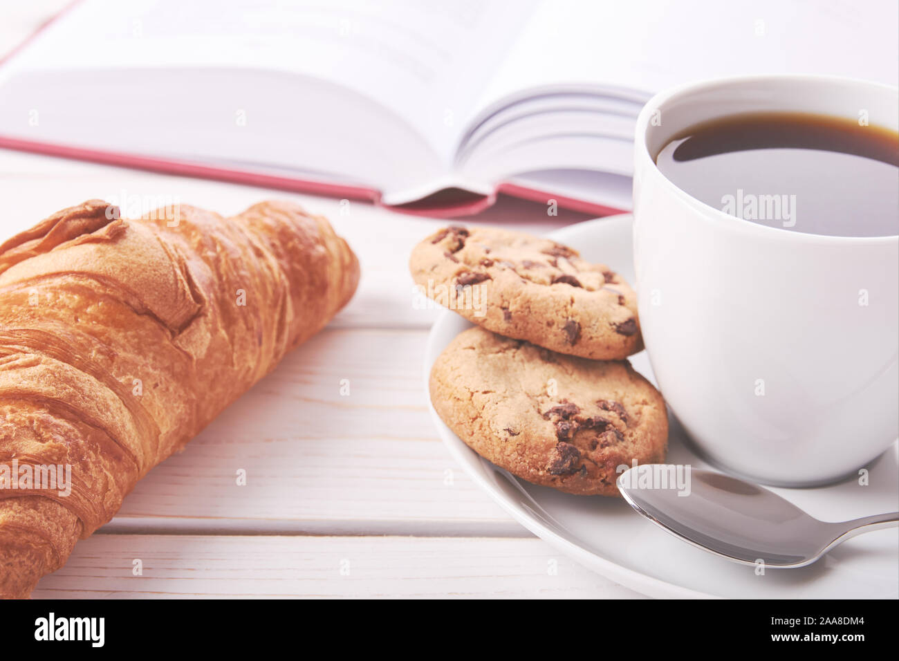 Still life with red book, cup of coffee or tea and two cookies. Croissant on white table top. Bright photo of breakfast. Stock Photo