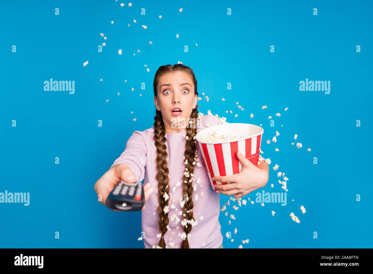 Portrait of afraid youngster girl with braids pigtails watch tv hold box with popcorn see horror series want switch channel pop corn flying falling Stock Photo