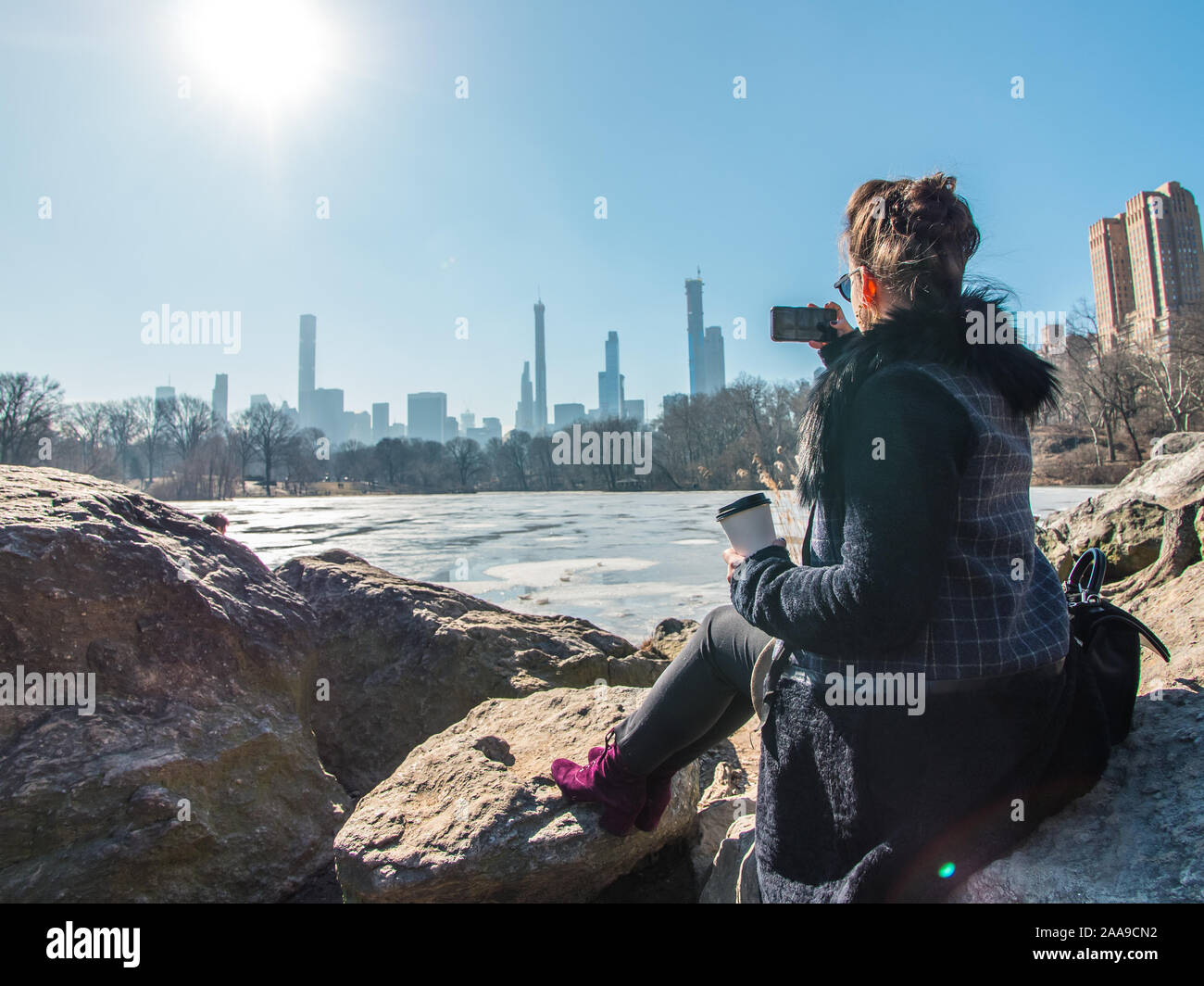 Young woman sitting on rocks by frozen lake in Central Park taking pictures of Manhattan Stock Photo