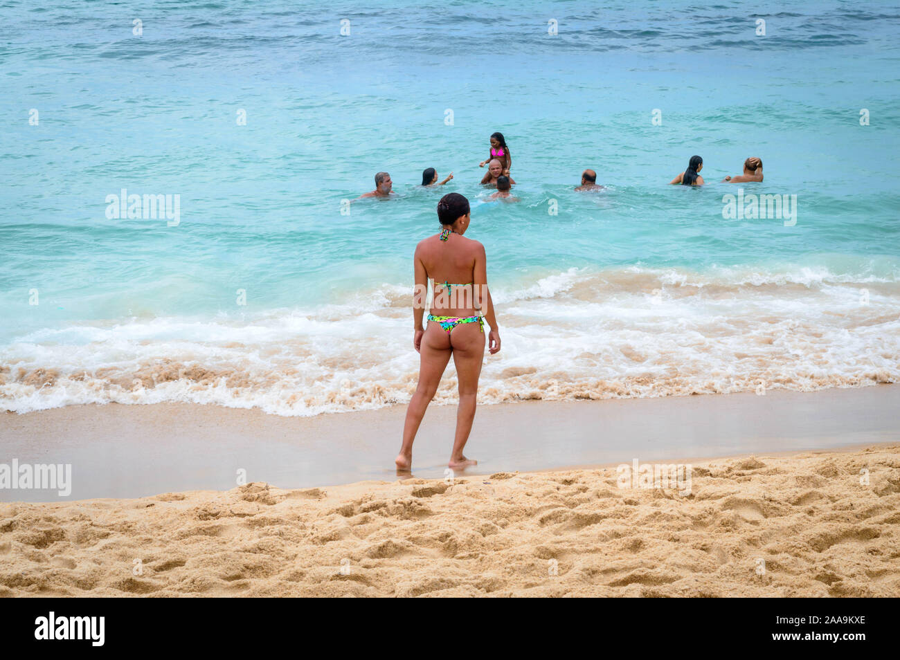 A Brazilian woman wearing a small bikini stands by the water watching the people who are bathing in the ocean. Stock Photo