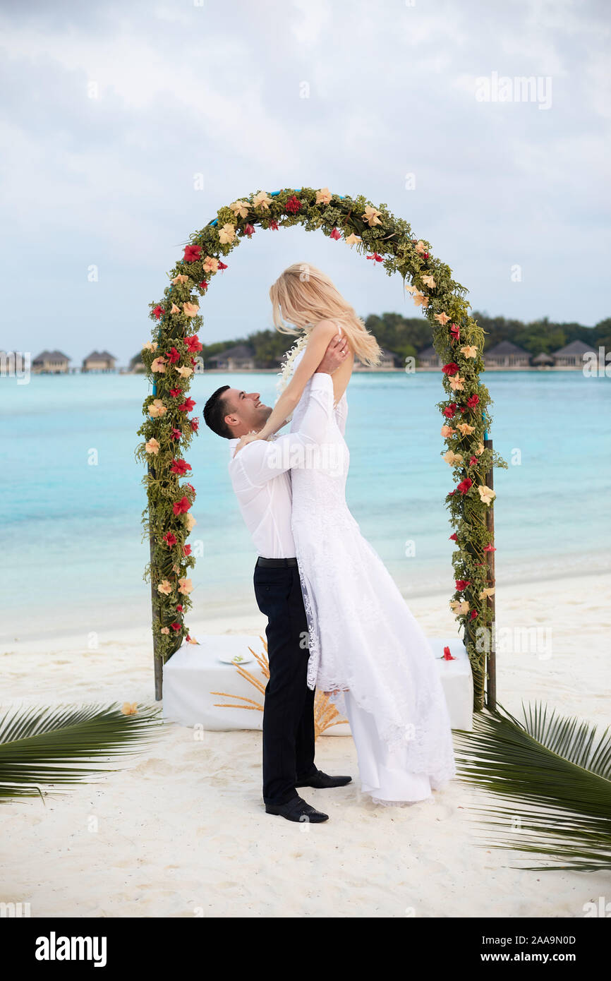 Happy groom holds bride on his hands under wedding ceremony arch on  tropical island beach on Maldives. Turquoise ocean lagoon, white sand and  Stock Photo