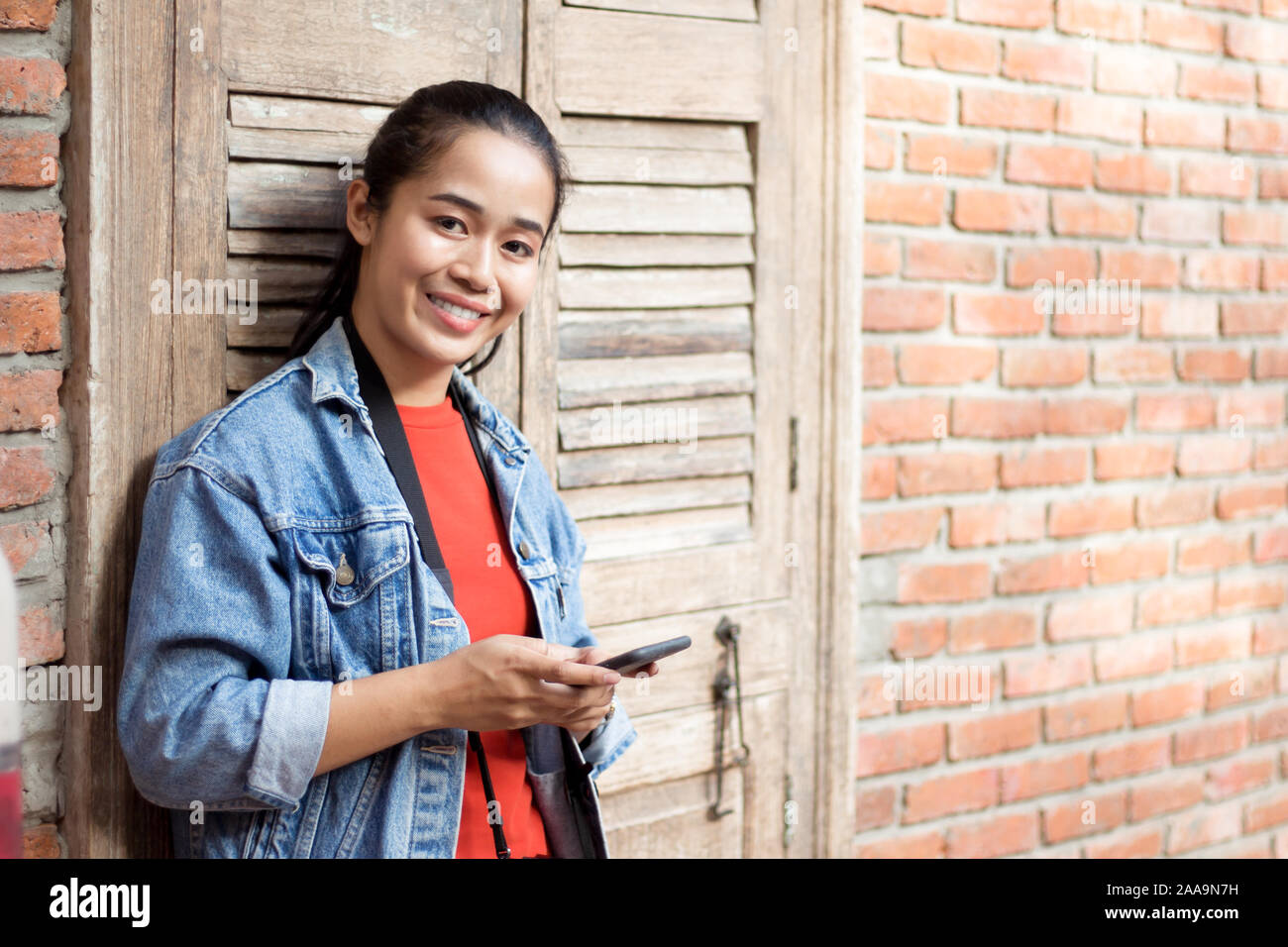 Teenage girl wearing a jacket, jeans and orange shirt standing against a wall and holding the phone with a smile. Stock Photo