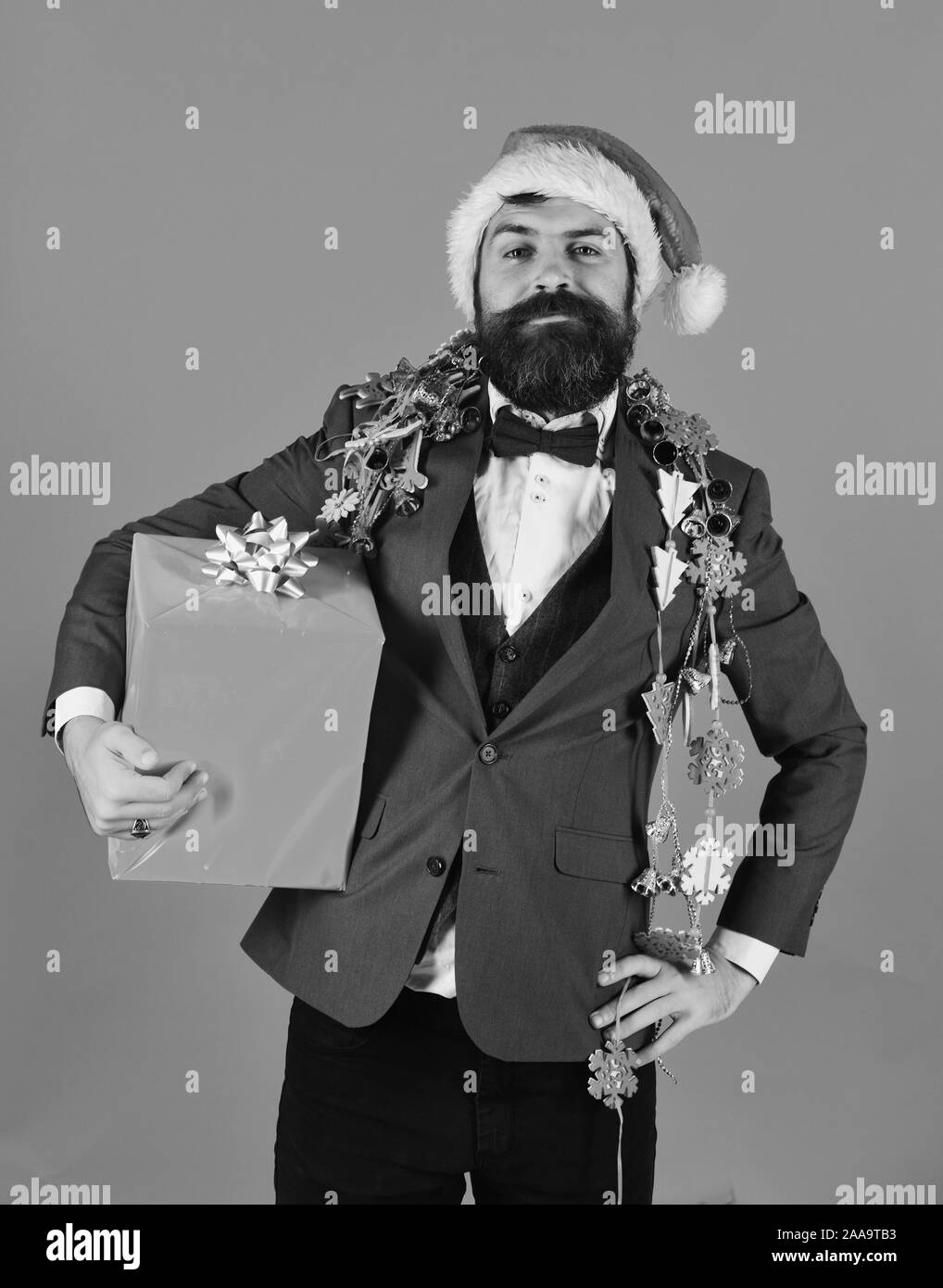 Manager with beard holds red gift. Man in smart suit, Santa hat and garlands on blue background. Businessman with proud face holds present box and decorations. Business and celebration concept. Stock Photo