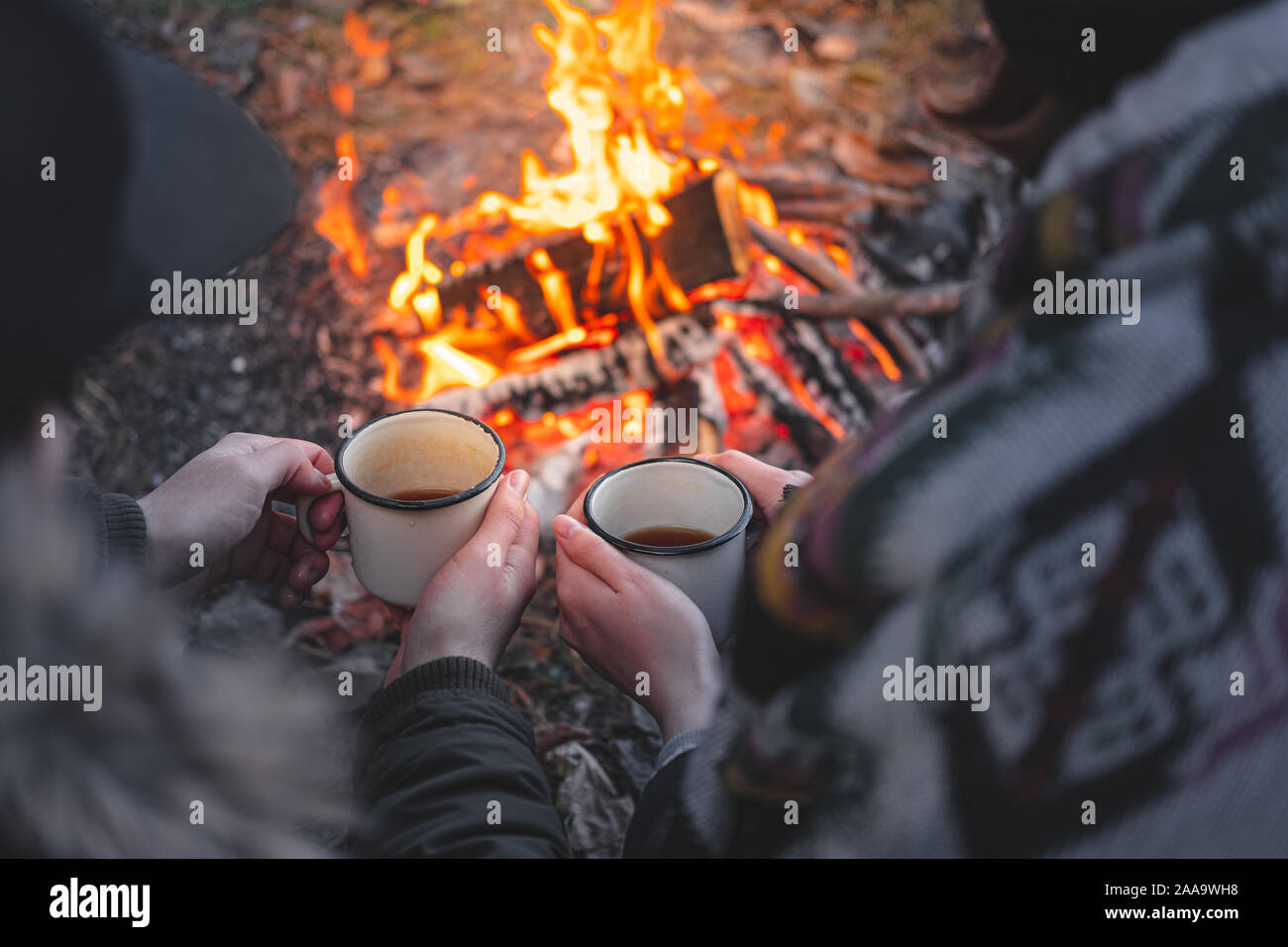 Two people warming hands with hot drinks by the bonfire. Spending nice time outdoors in chilly weather at a camping place - tranquil and peaceful scen Stock Photo