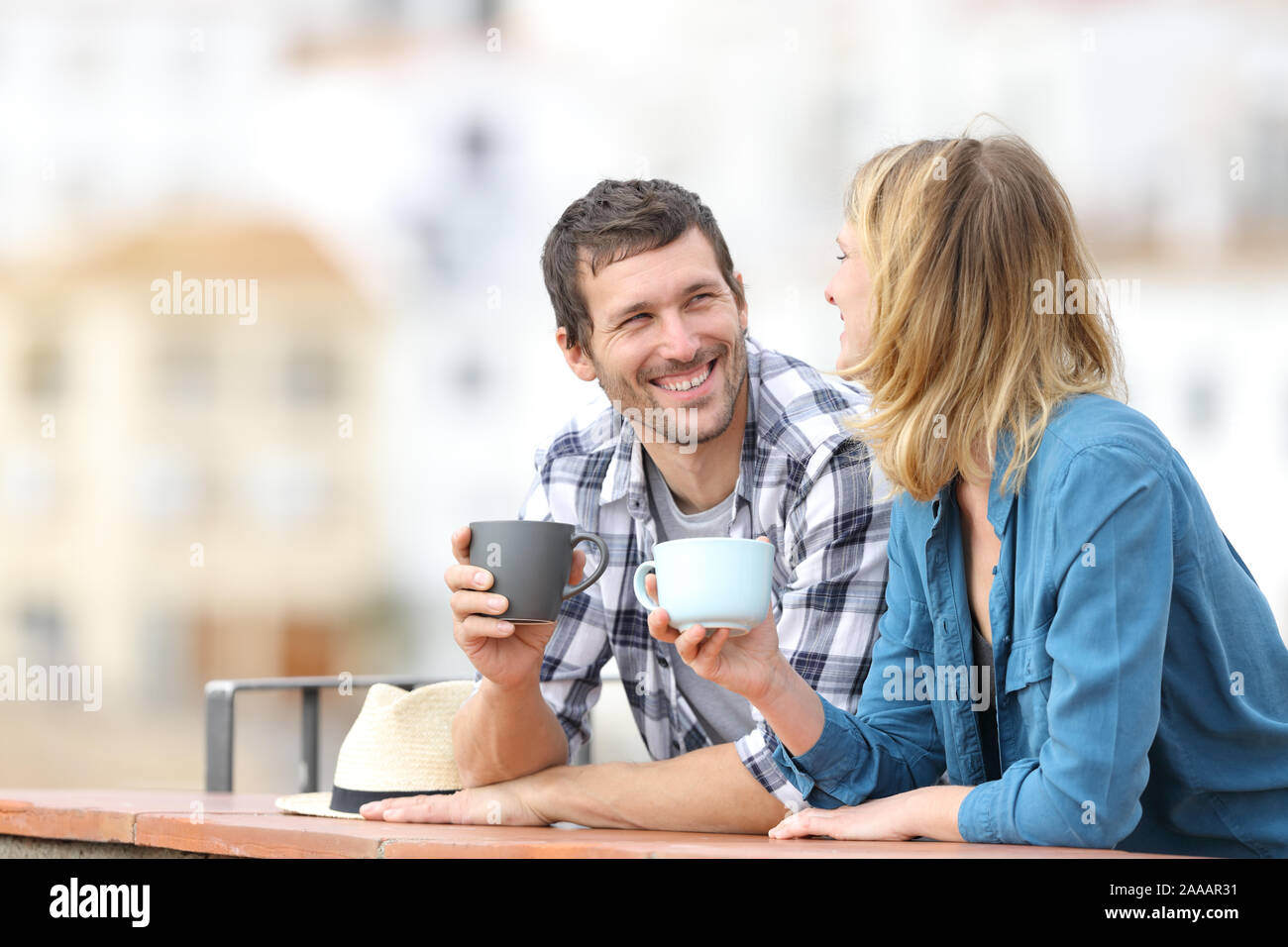 Happy couple talking holding coffee mugs standing in a balcony outdoors in a rural town Stock Photo