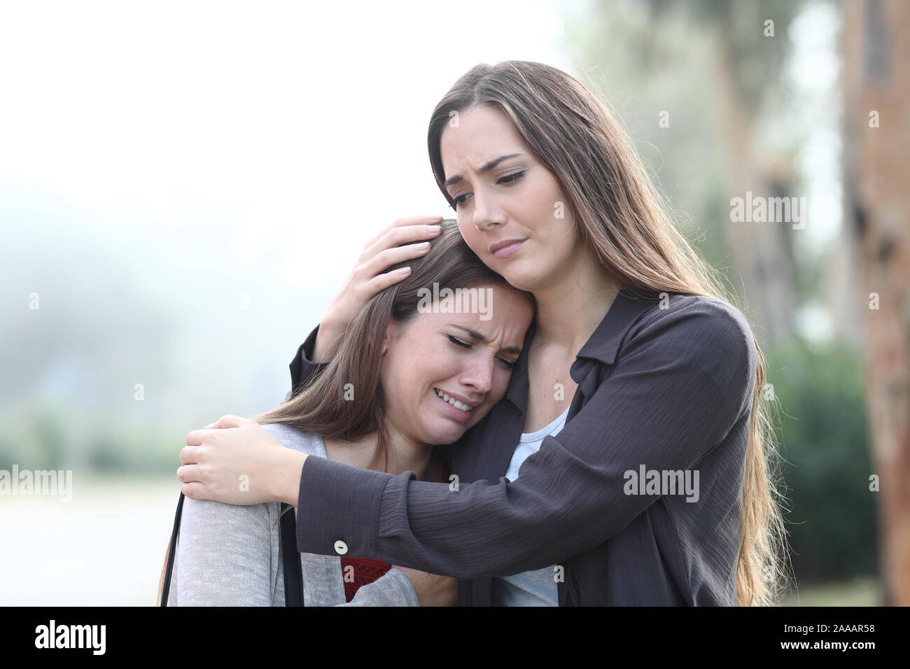 Sad woman crying and a friend comforting her standing in a park a foggy day Stock Photo