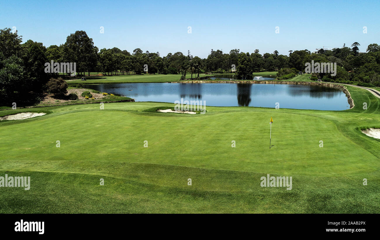 Golf course fairway, water hazard pond, sand bunkers, green with flag, lush green grass surrounded by trees against blue sky Stock Photo