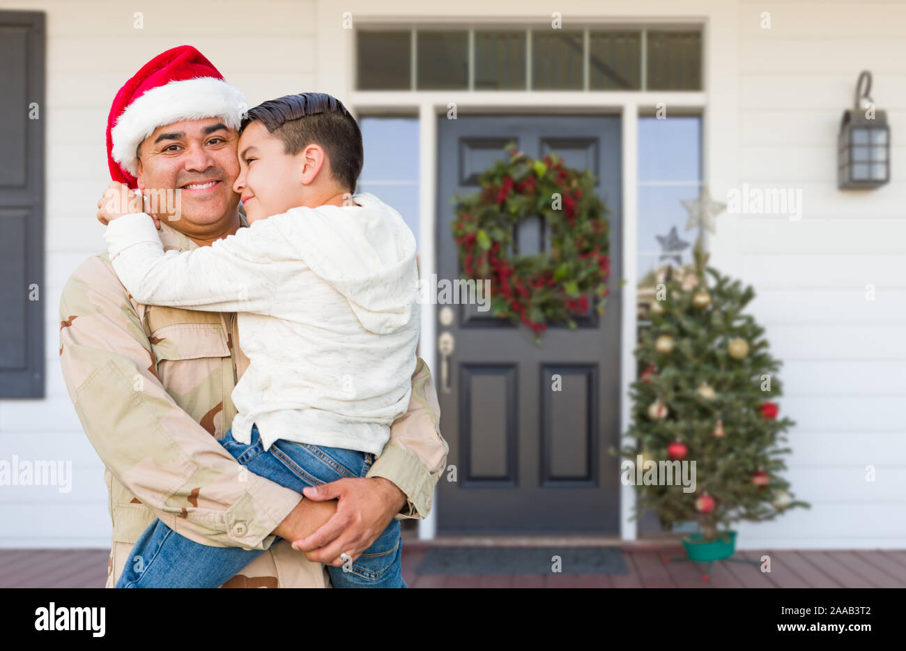 Hispanic Male Soldier Wearing Santa Cap Holding Mixed Race Son In Front of House. Stock Photo
