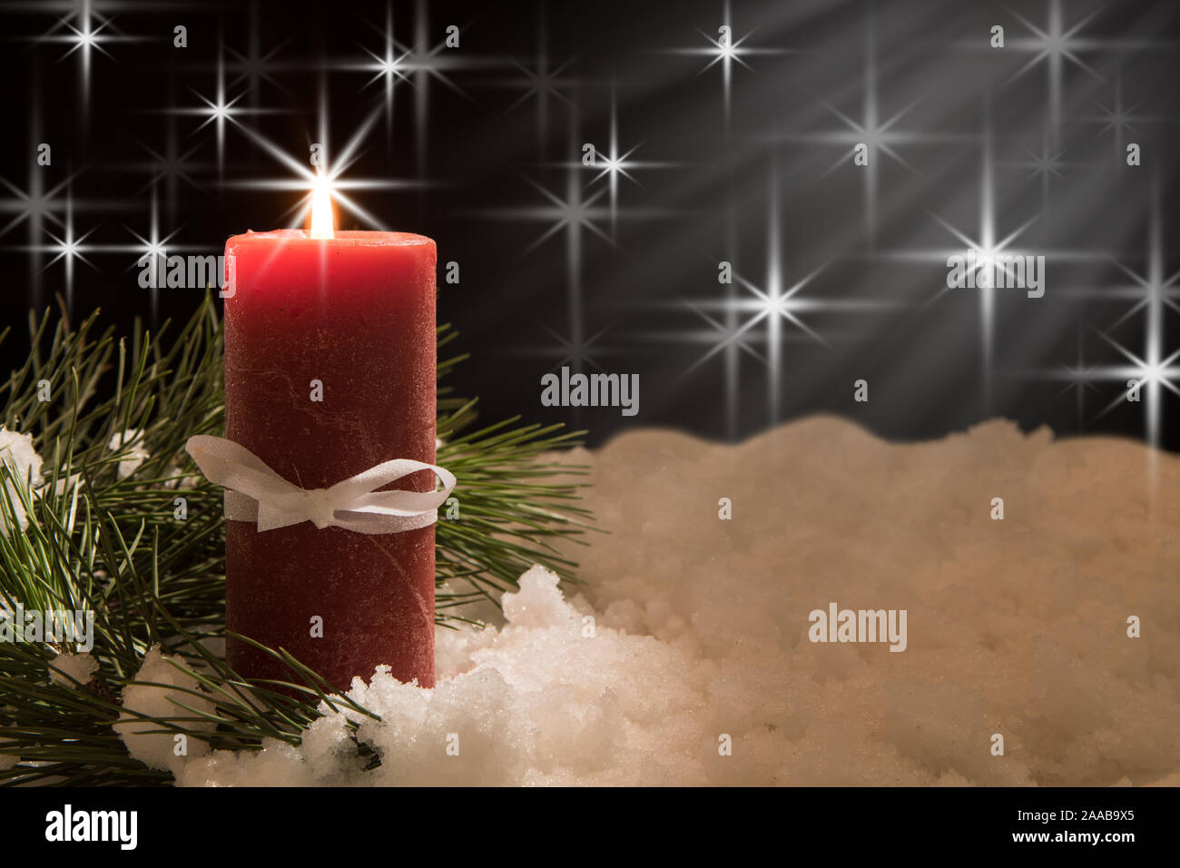 lit white candle and red ribbon with pine needles with beam of light in snow. Black Christmas Background with holiday theme. Stock Photo
