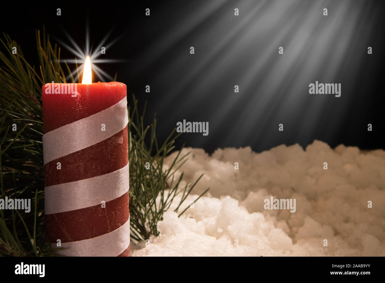 lit red candle and white ribbon with pine needles with beam of light in snow. Black Christmas Background with holiday theme. Stock Photo