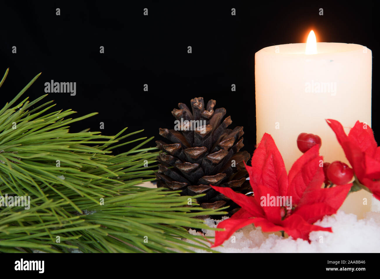 Red Poinsettia flower with lit white candle, pine needles and pine cones in snow. Black Christmas Background with holiday theme. Stock Photo