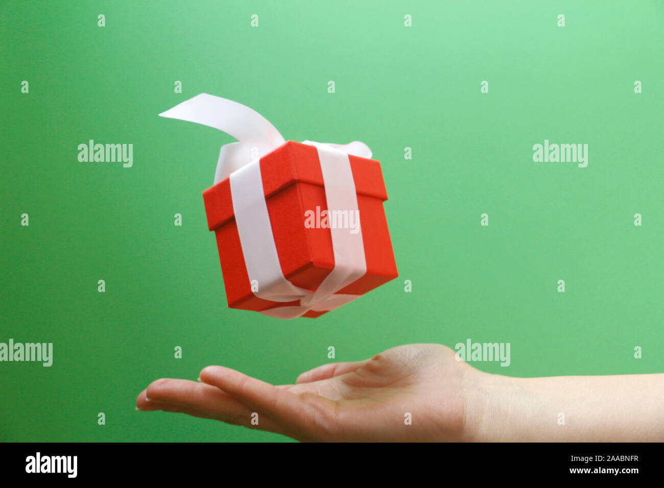 Red gift box with a white bow on a green background. The box is closed by a lid. A gift flies in the air. Under the gift is a female hand. Gift levita Stock Photo