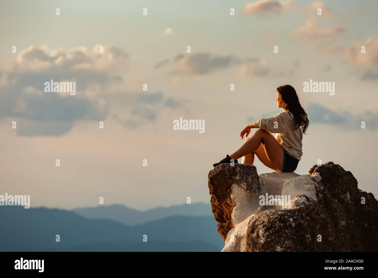 One sporty girl sits on big rock and looks at sunset sky and mountains Stock Photo