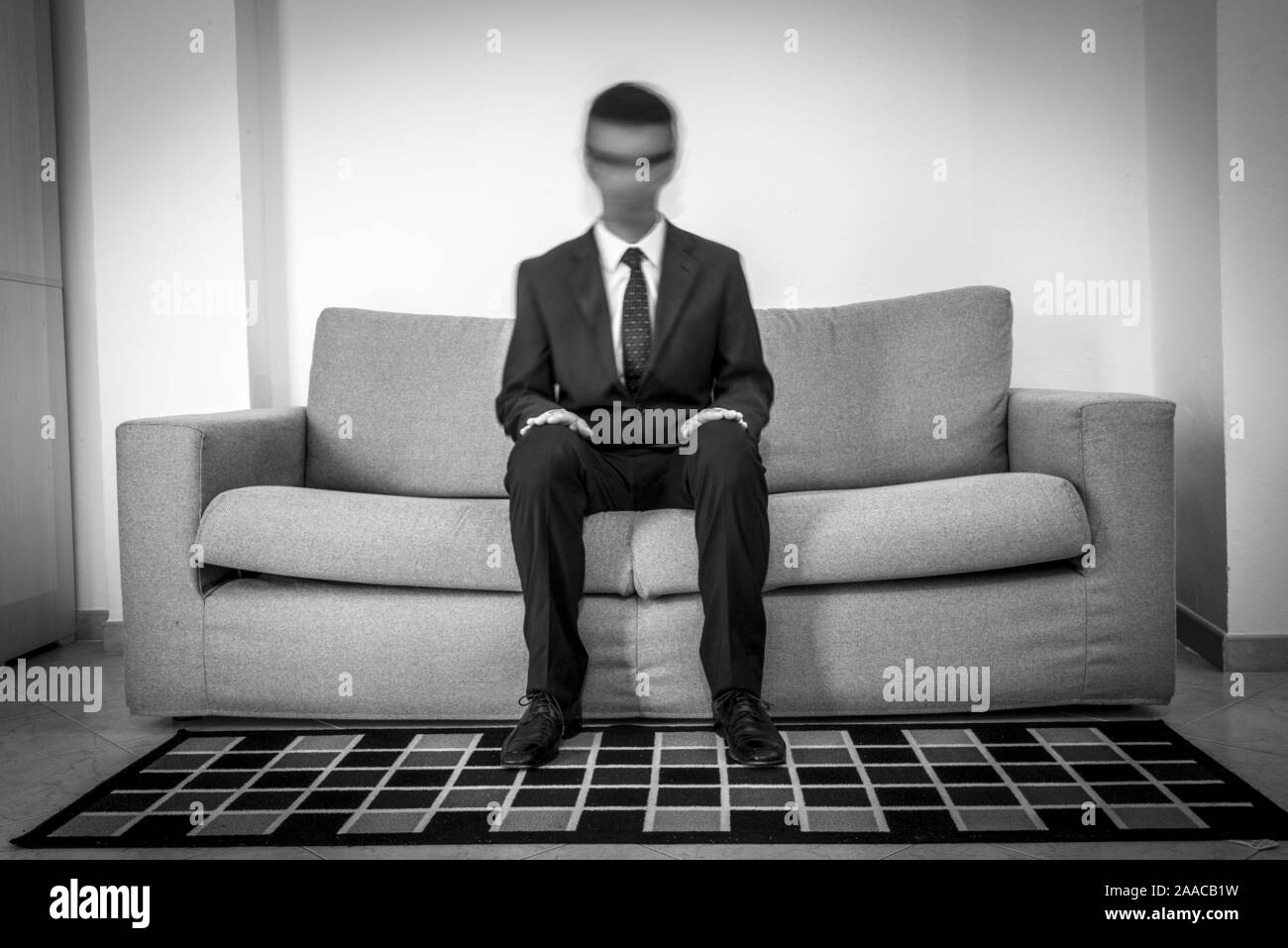 Man in suit and tie with blurred face sitting alone in the sofa. Concept of mental illness and depersonalization. Black and white Stock Photo
