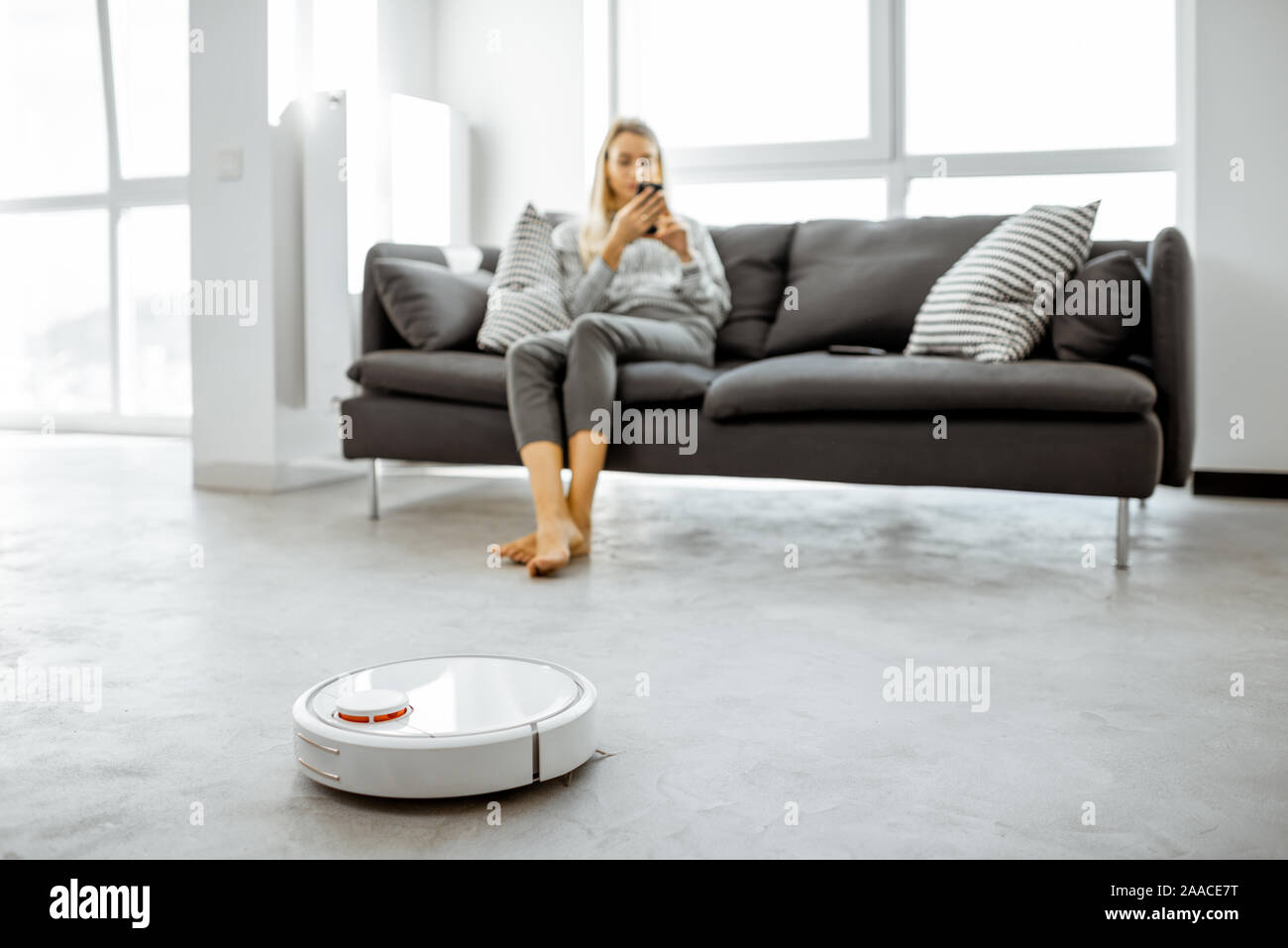 Woman relaxing on the couch while automatic vacuum cleaner doing the housework in the modern white living room. Household robots concept Stock Photo