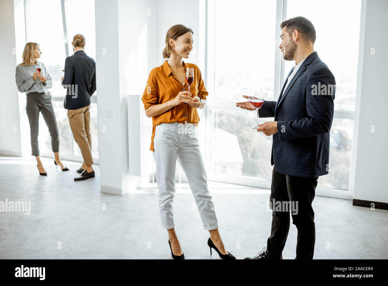 Young elegantly dressed people meeting in the white hallway or showroom, talking and drinking wine during some informal event Stock Photo