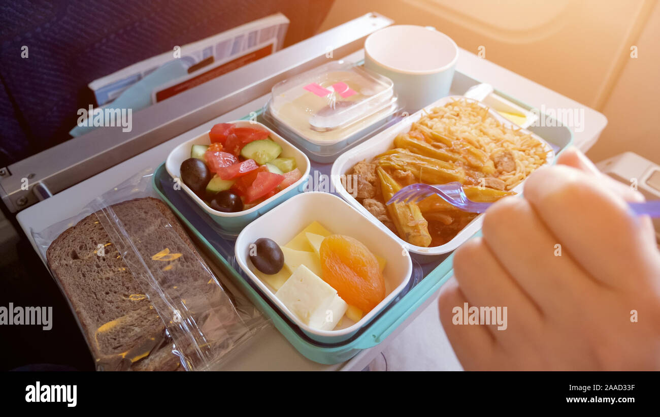 Lunch in plane: bread, meat with vegetables and rice, pieces of cheese,  dried apricot, salad and beverage in cup. Woman is eating in airplane  during flight food served on tray, hand with