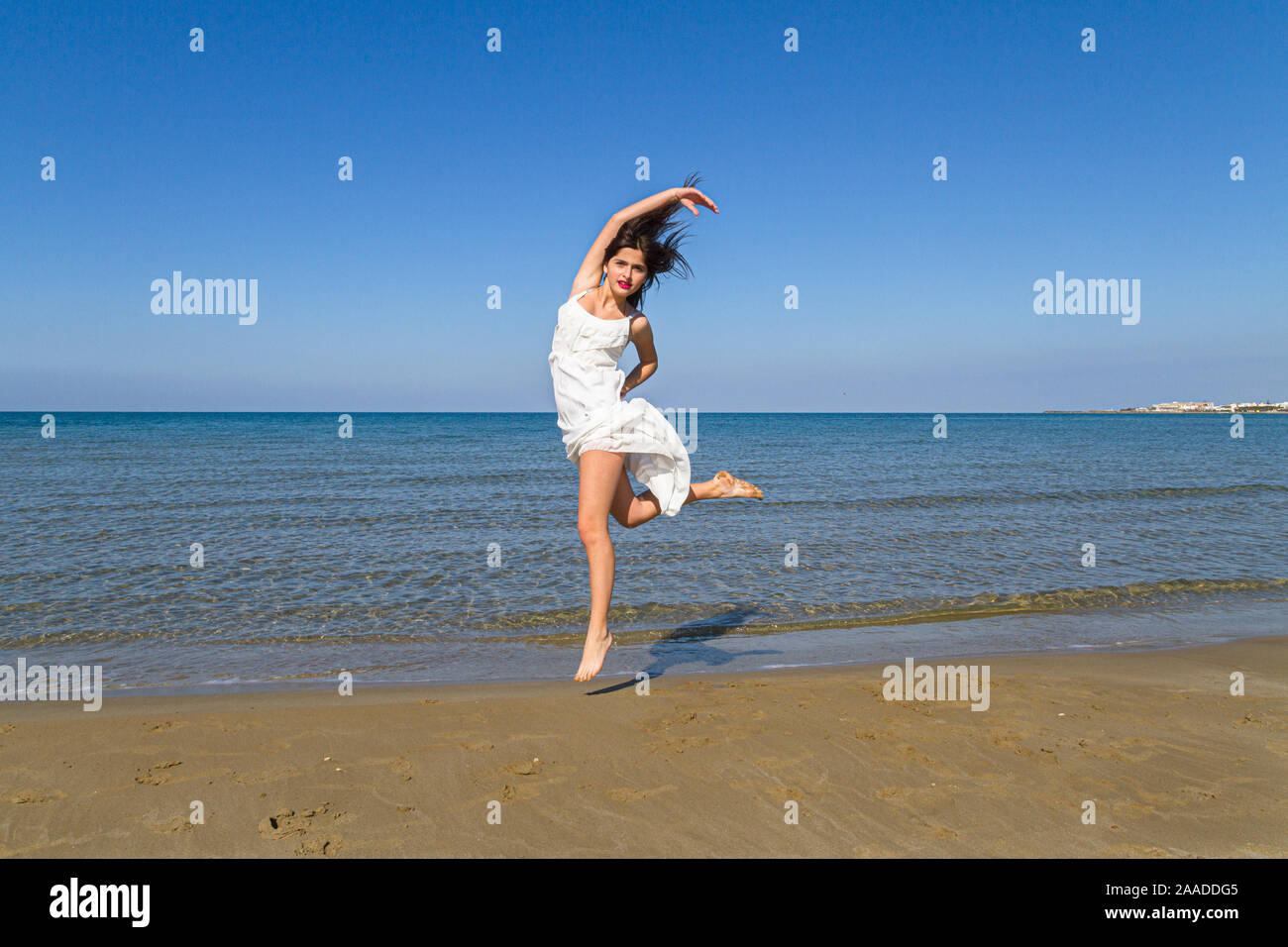 Young carefree beautiful girl in flimsy white dress. Fresh young beautiful joyful woman jumping on the beach in summer. Stock Photo