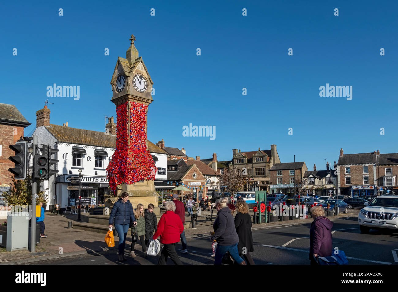 People using pelican crossing and Market Square clock decorated with knitted poppies for Remembrance Day Thirsk North Yorkshire England UK United King Stock Photo