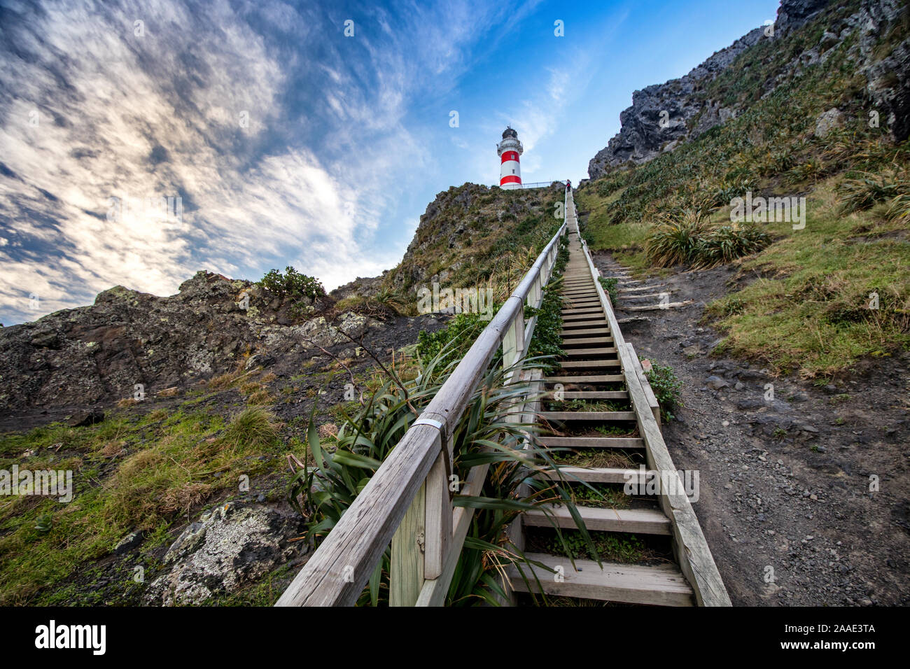 View on Cape Palliser red and white striped lighthouse with steep wooden stairs leading to it. Lighthouse sits on rocky ridge with lush vegetation. Stock Photo