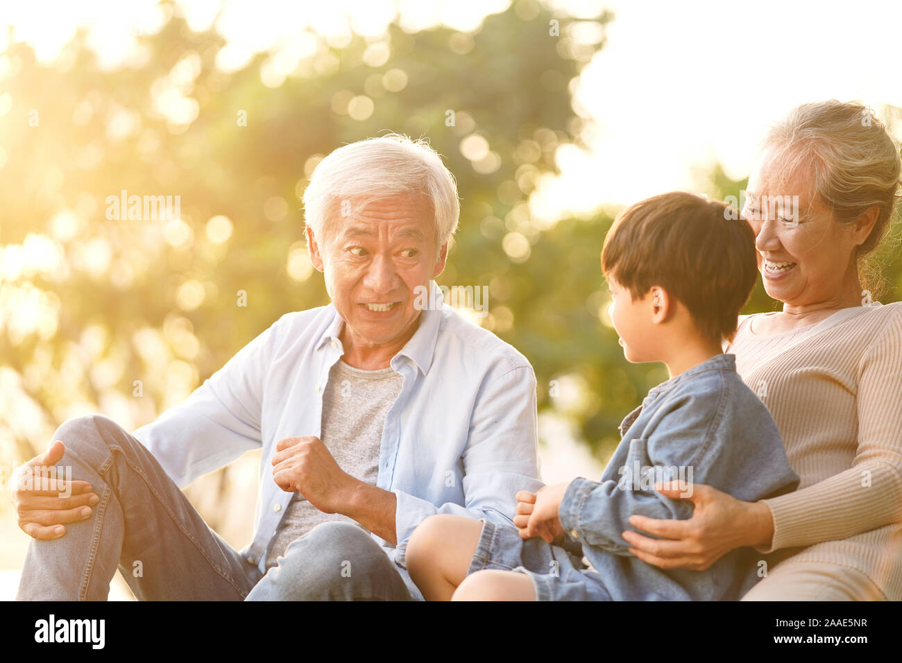 asian grandson, grandfather and grandmother sitting chatting on grass outdoors in park at dusk Stock Photo