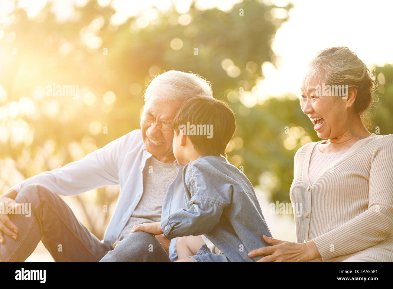 asian grandson, grandfather and grandmother sitting on grass having fun outdoors in park at sunset Stock Photo