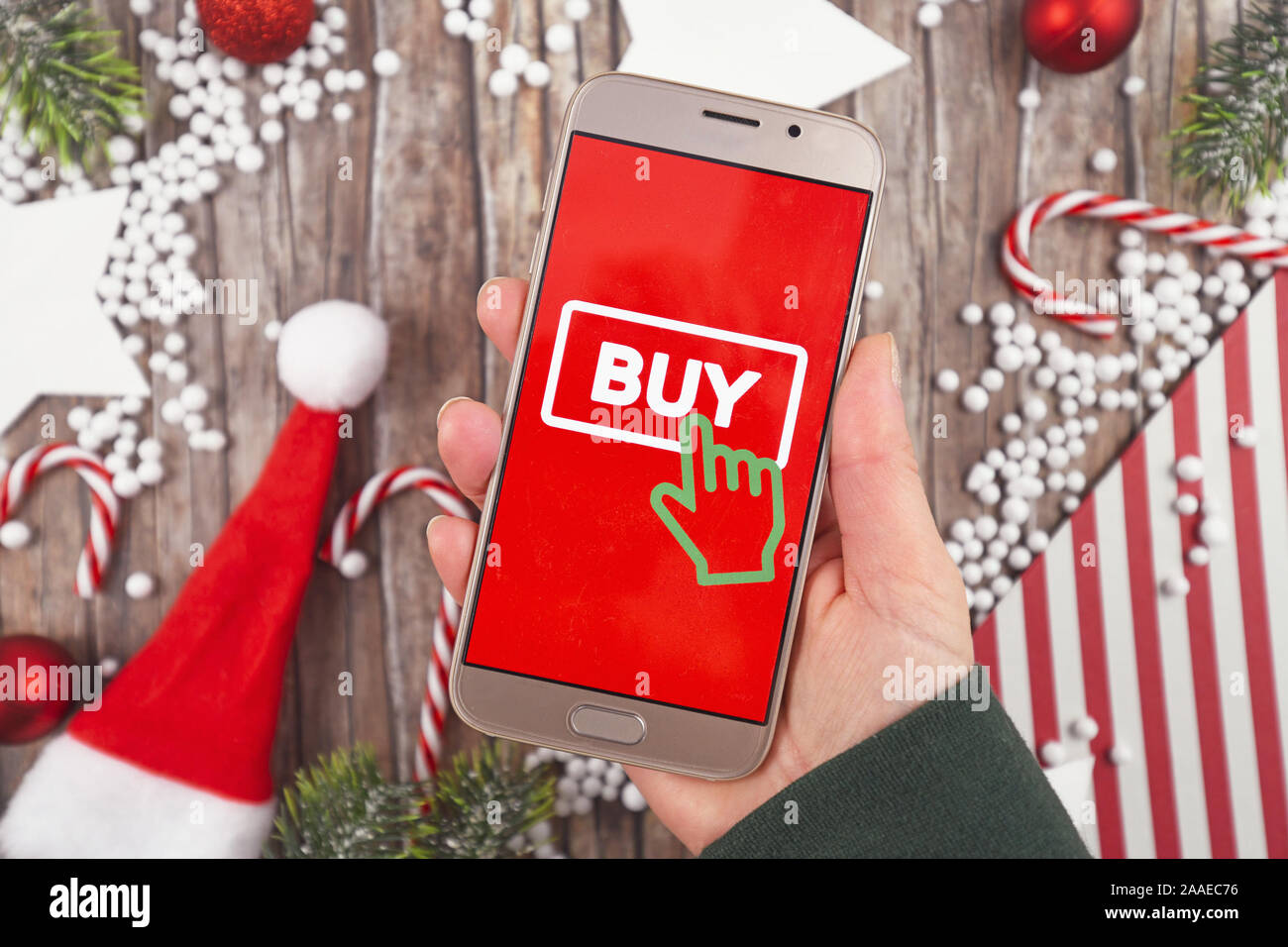 Concept for Christmas seasonal online shopping and sales with hand holding cell phone with 'Buy' sign button in red background in front of wooden desk Stock Photo