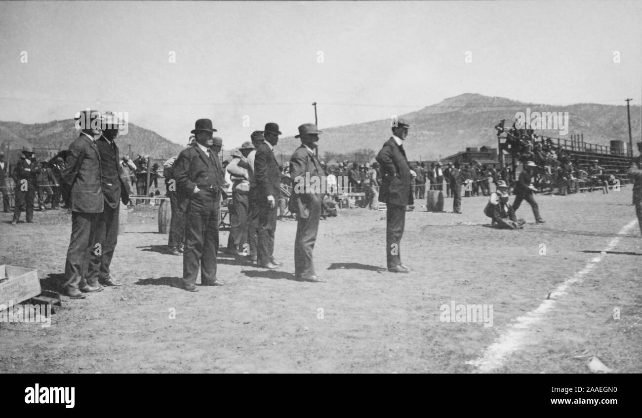 Full-length profile view of several men, wearing suits and hats, standing at the edge of a crowded field on a sunny day, watching a shooting contest taking place off-camera, New Mexico, 1910. () Stock Photo