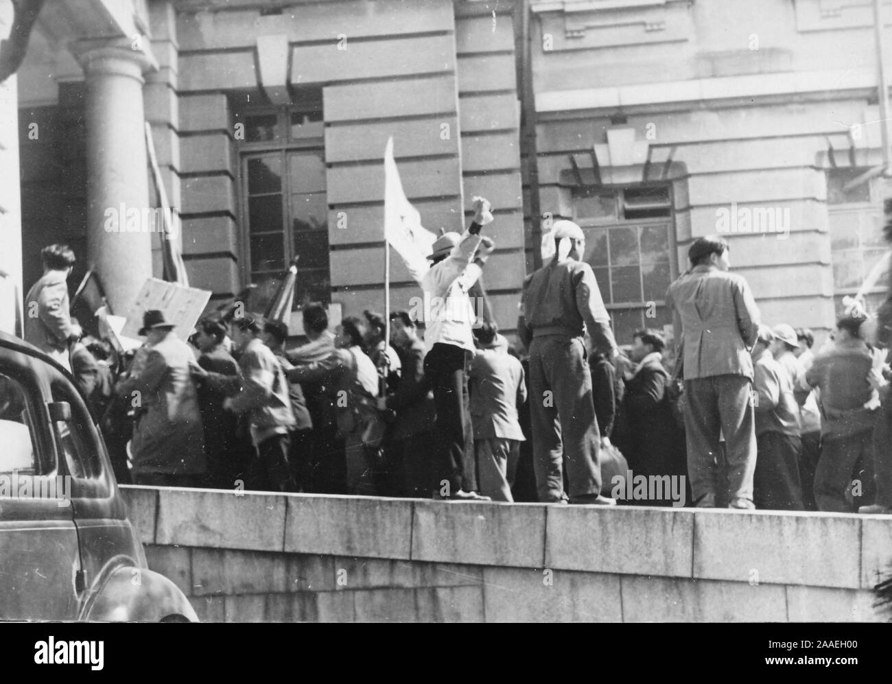 A crowd of Japanese workers, from the back and in profile, hold flags, stand on a wall, and march towards a building entrance, likely the old Fukuoka Prefectural Office, Fukuoka Prefecture, Japan, 1950. () Stock Photo