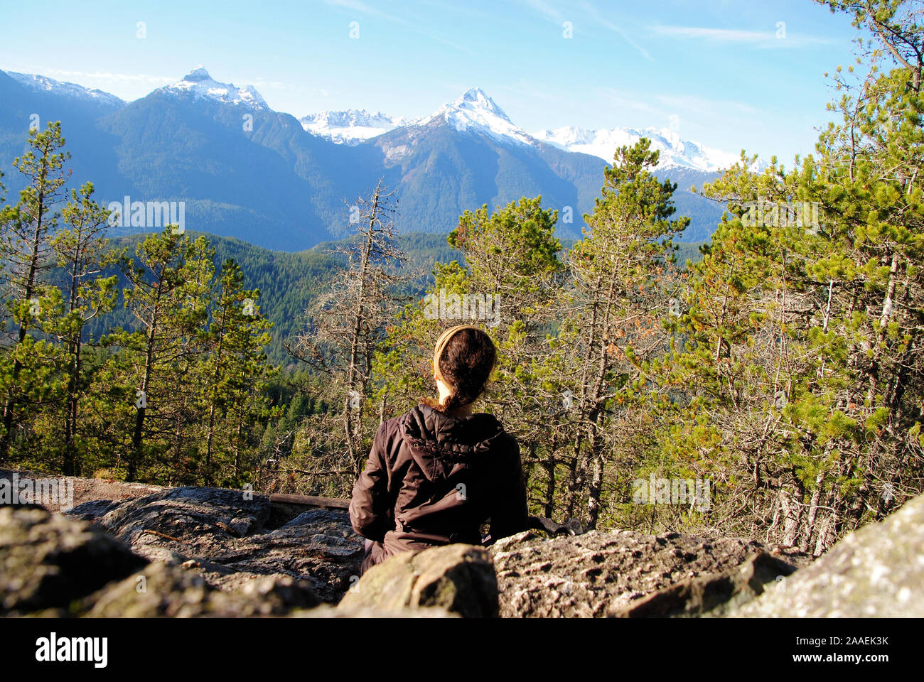 A natural young woman with brown braided hair, sitting still on rocks in a contemplative posture, staring at the snow capped Tantalus mountain range Stock Photo