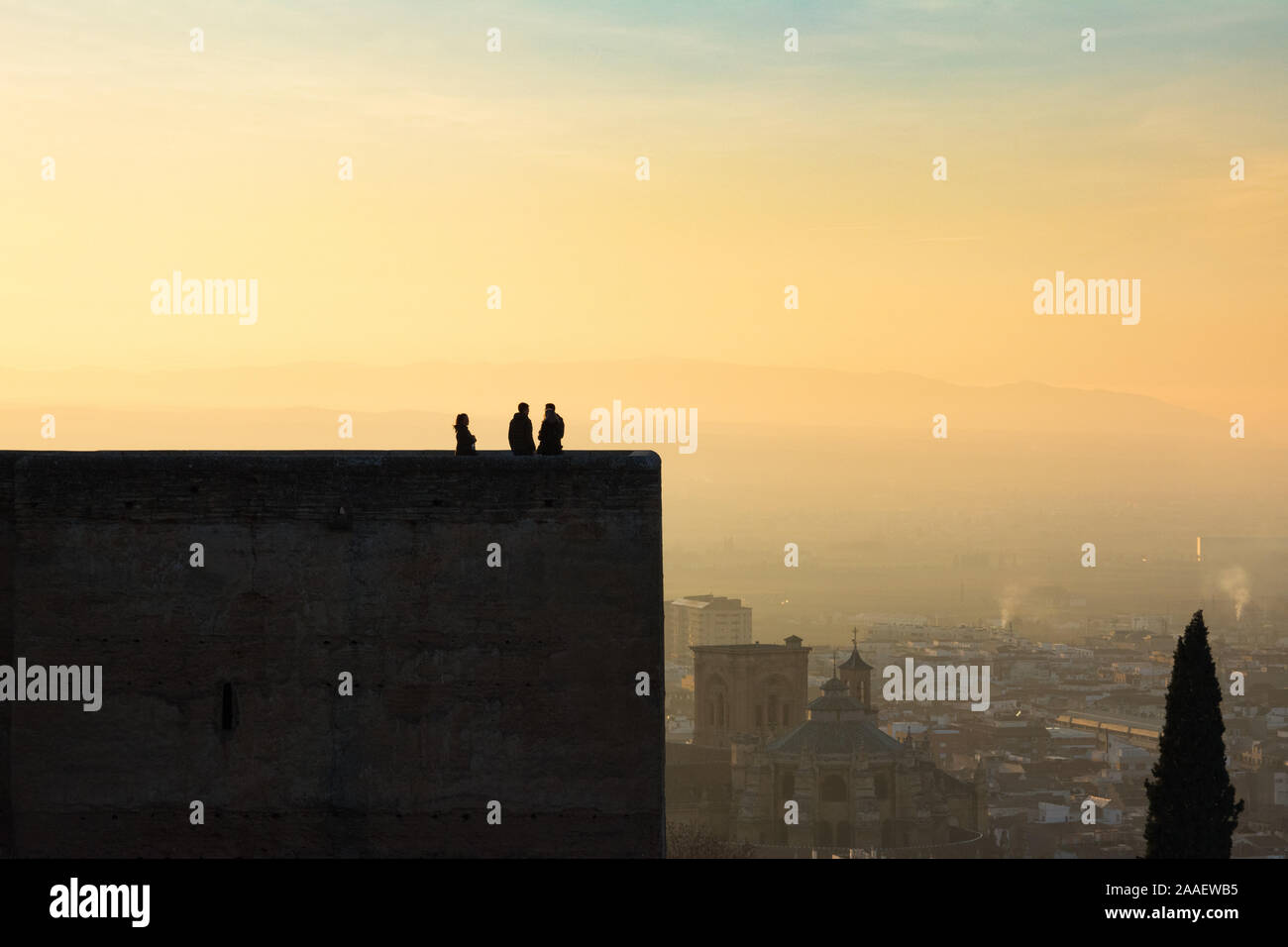 People gaze at the Spanish city of Granada from the Alcazaba fortress of the Alhambra palace during sunset. Grenada Cathedral can be seen beyond. Stock Photo