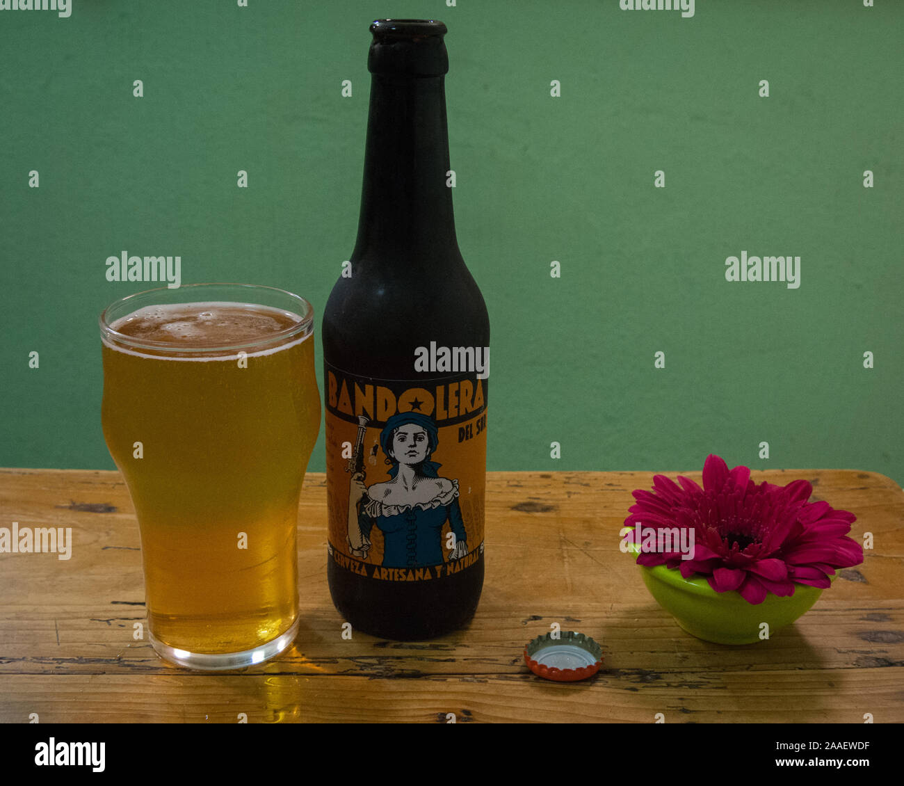 A wooden table with an opened bottle and full glass of Spanish artisanal pale ale called Bandolera, and a small flower vase. Stock Photo