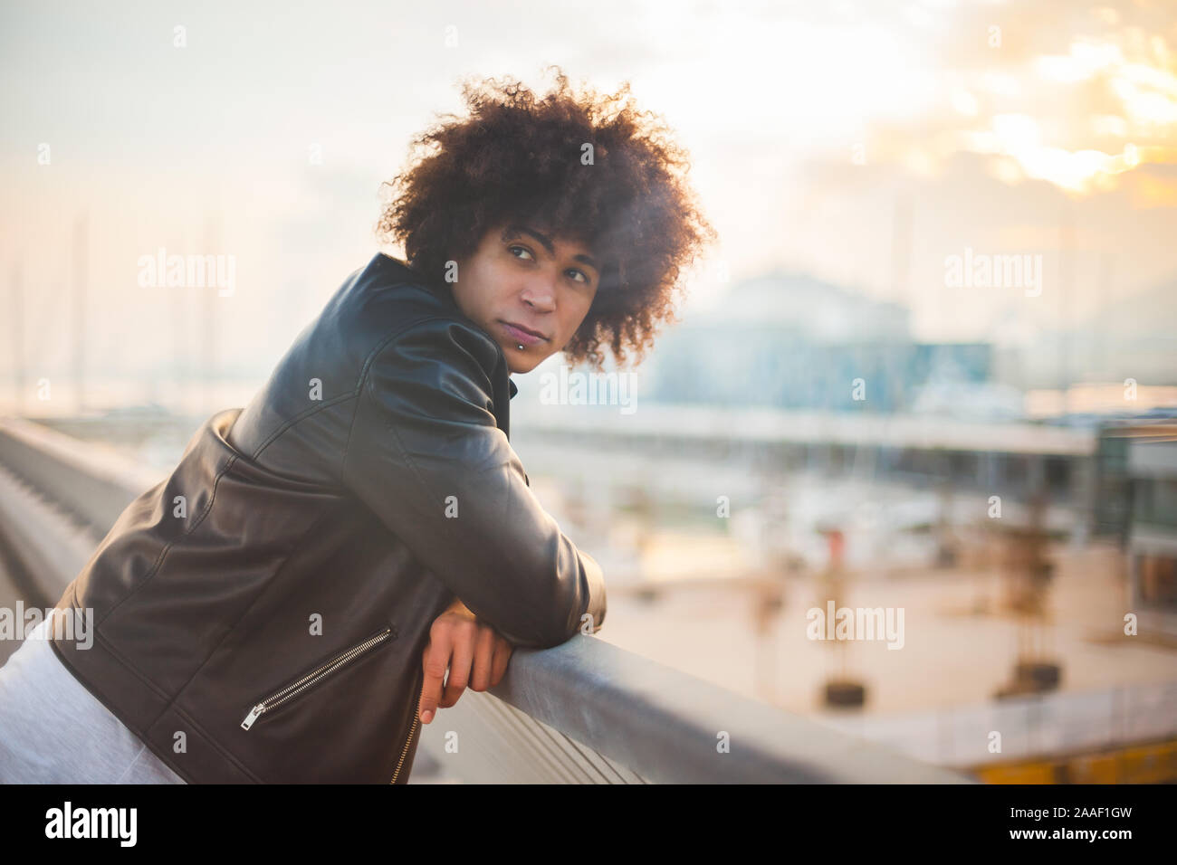 Young mixed-race man with afro hairstyle standing at a handrail with beautiful sunset with urban background Stock Photo