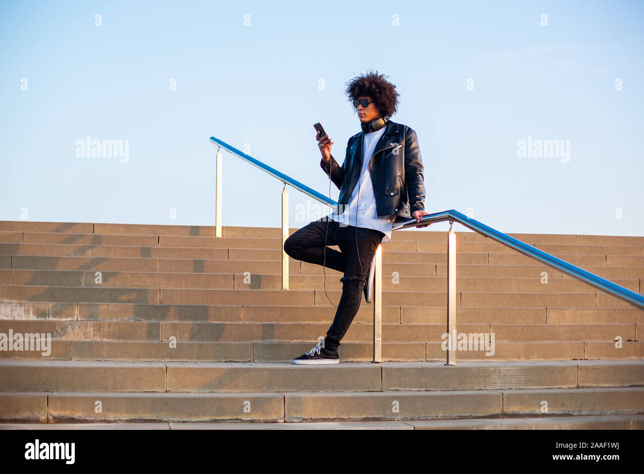 Handsome young man with afro hairstyle standing on stairs using a smart phone outdoors with blue sky Stock Photo