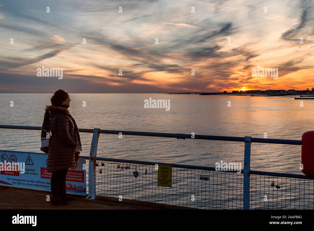 A woman looks out across the sea to the sun setting behind a coastal town. Concept of sun setting on a relationship, falling out of love, breaking up. Stock Photo